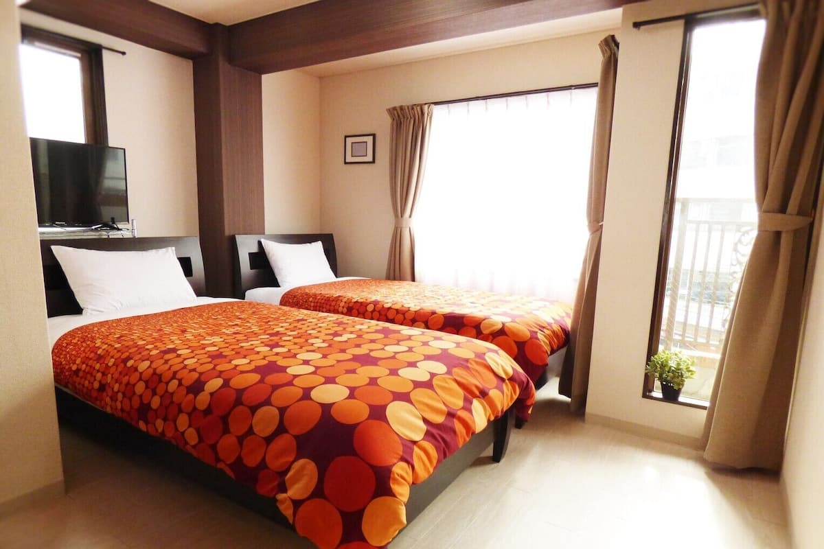 A hotel room with two beds, a TV, a window with curtains, and a balcony with a potted plant.