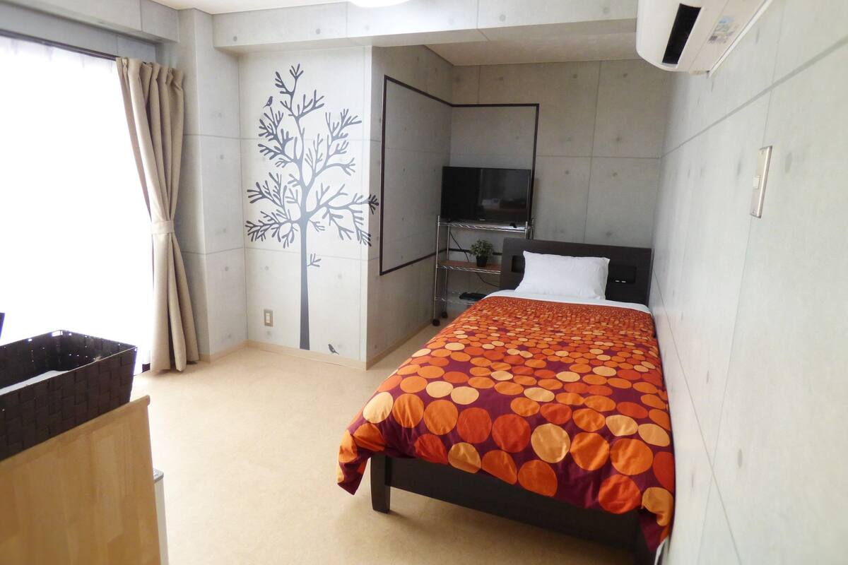 A hotel room with a bed, a TV, a wall-mounted shelf, and a window with curtains.