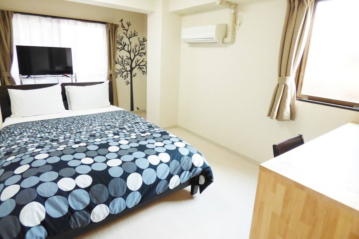 A hotel room with a bed, a TV, an air conditioning unit, a desk, and a window with curtains.