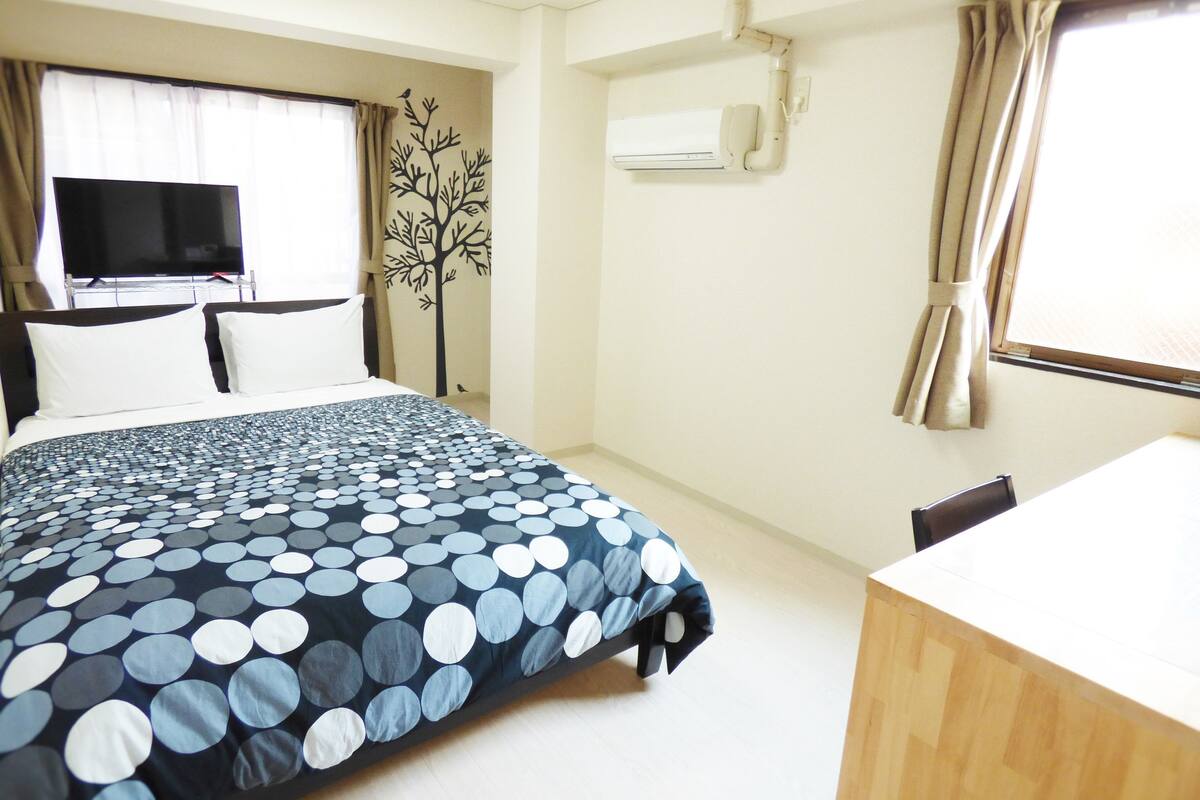 A hotel room with a bed, a TV, a desk, and a window with curtains.
