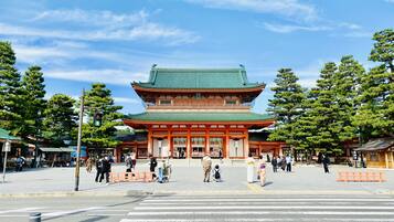 Un bâtiment traditionnel japonais, doté d’un toit végétalisé et de piliers rouges, entouré d’arbres et bordé par un passage piéton.