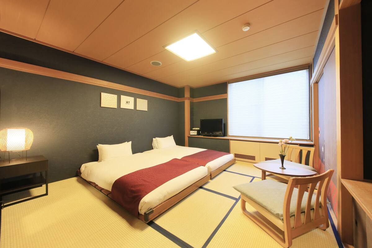 A hotel room with a bed, a bedside table with a lamp, a small table with a vase, and a window with blinds.
