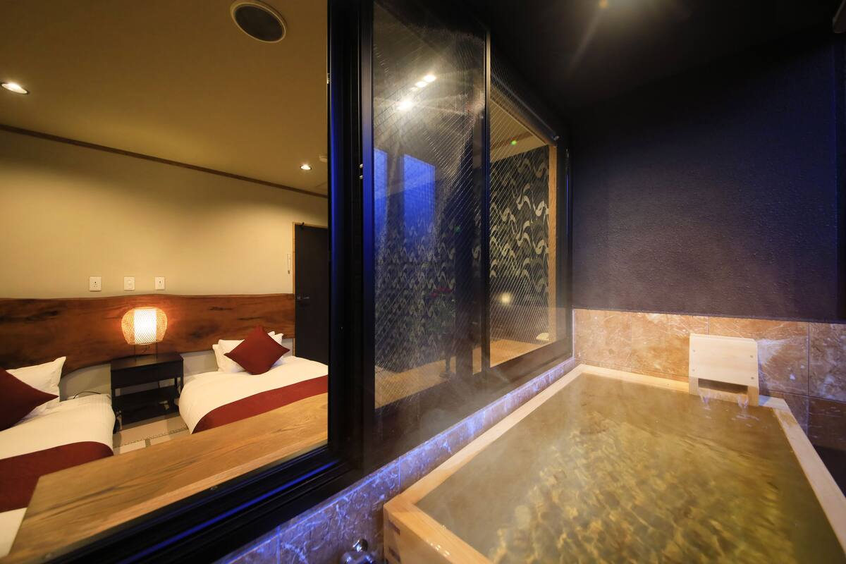 A modern hotel room with a large glass window, a wooden headboard, a small bedside table with a lamp, a large bathtub, and a view of the outside.