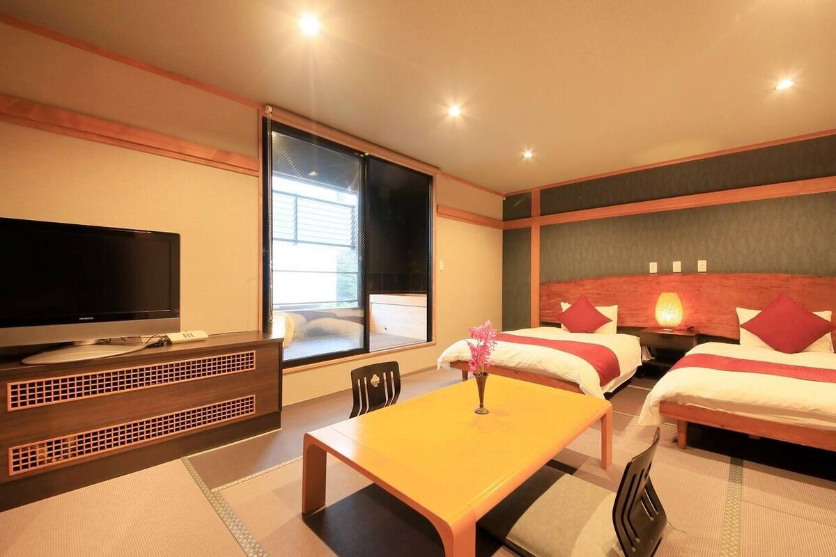 A hotel room with a television, a dining table, two beds, and a window.
