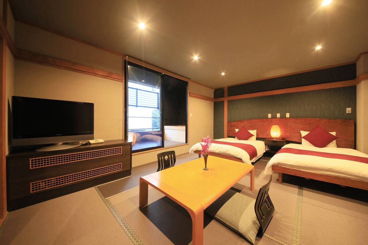 A hotel room with a television, two beds, a table, and a window with blinds.