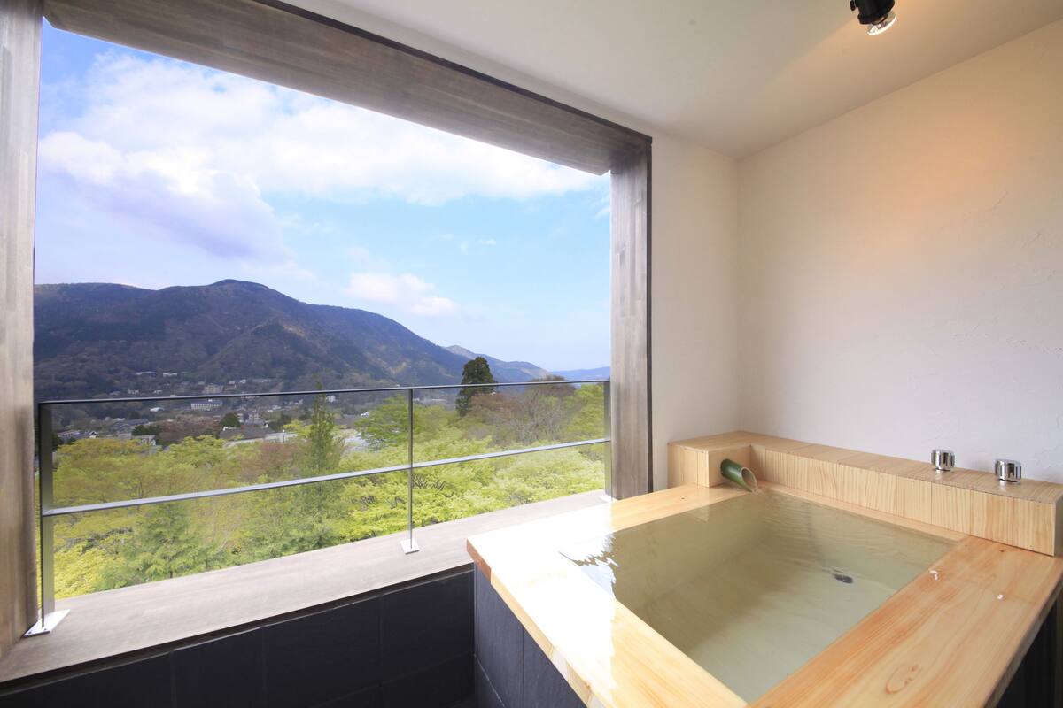A modern bathroom with a large wooden bathtub, a glass-enclosed shower area, and a balcony with a scenic mountain view.