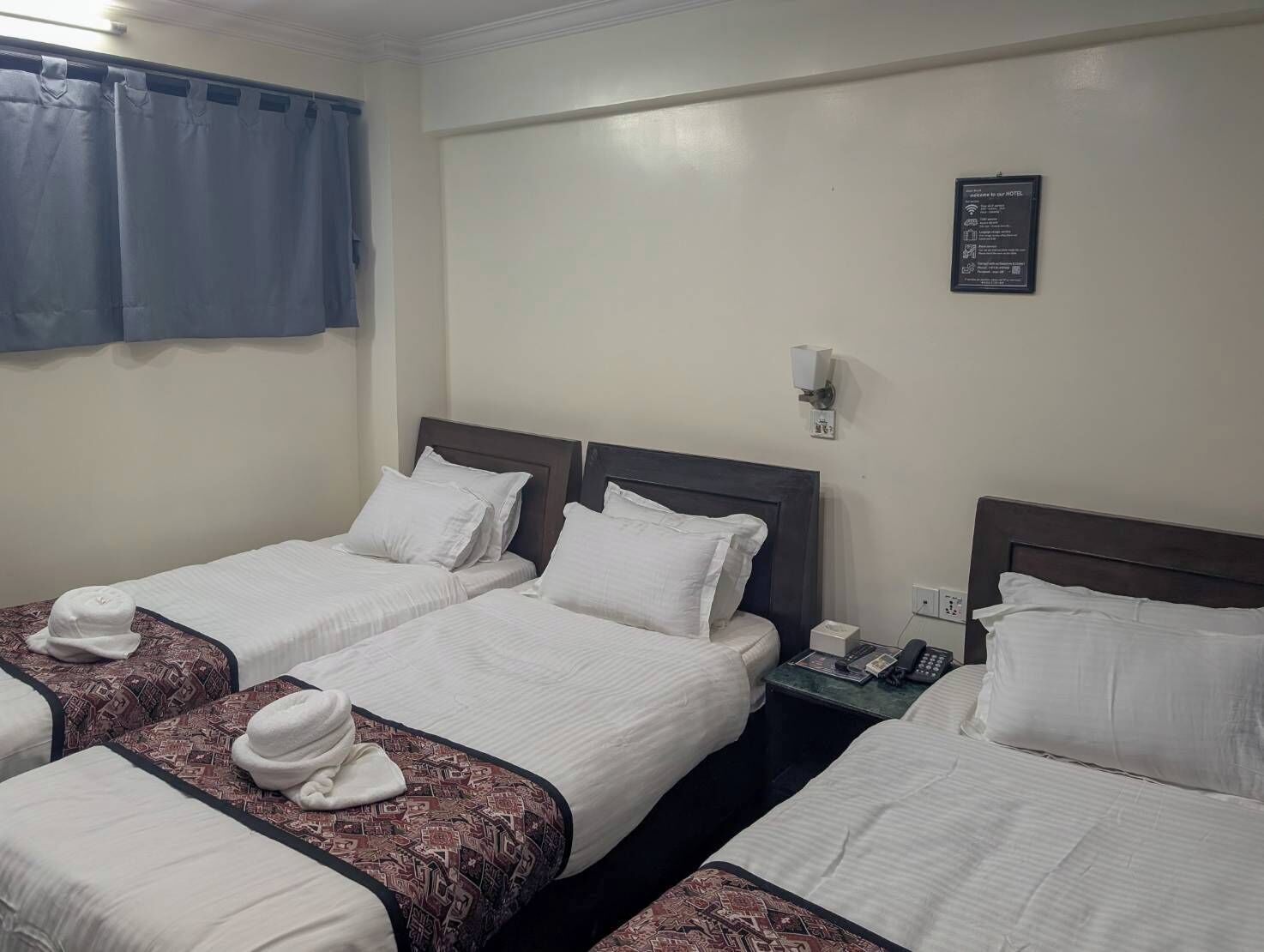 Een hotelkamer met drie eenpersoonsbedden, elk voorzien van witte lakens en een bedsprei met dessin. Er staat een nachtkastje met een telefoon en een wandlamp. Een blauw gordijn is gedeeltelijk opengetrokken, en een bordje is zichtbaar aan de muur.