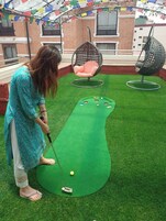 Una persona juega al golf sobre una alfombra verde, con dos sillas y un edificio al fondo.