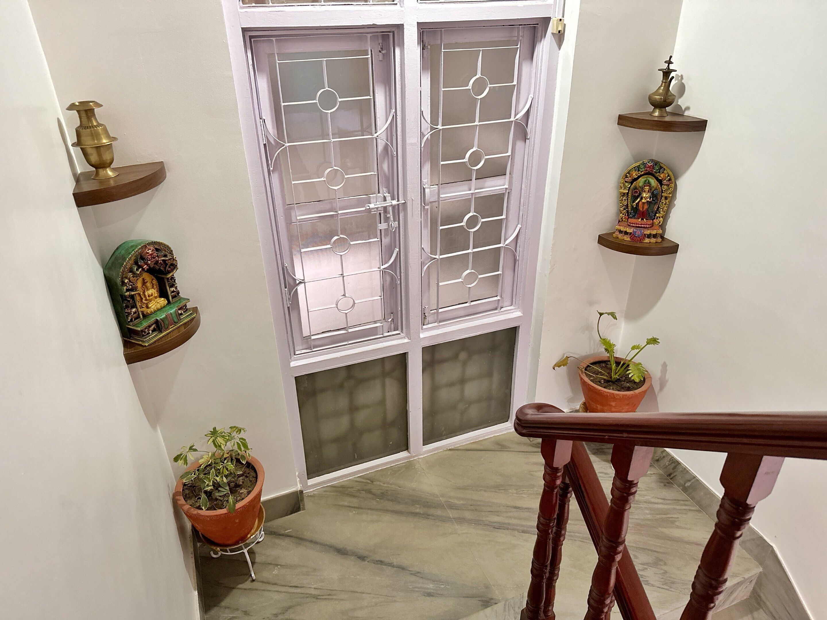 Una escalera con barandilla de madera, una puerta blanca con celosía metálica y plantas en macetas a ambos lados.