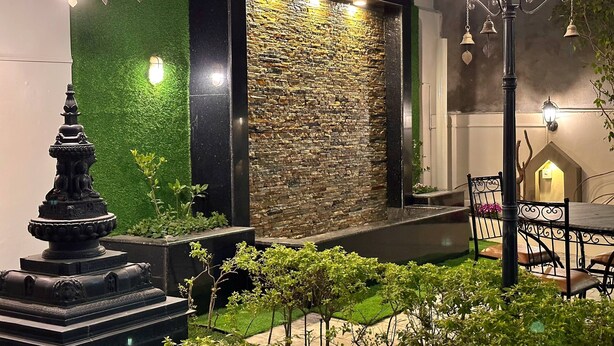 An outdoor seating area with a stone wall backdrop, a Buddha statue, and a lamp post. - Shakya House (Lalitpur)