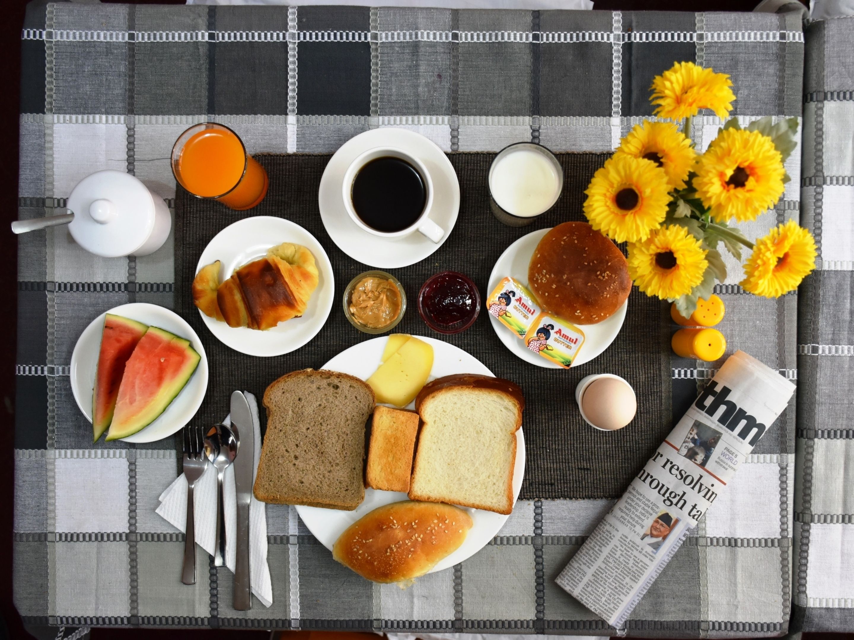 A breakfast table with various food items, a newspaper, and a vase of sunflowers.
