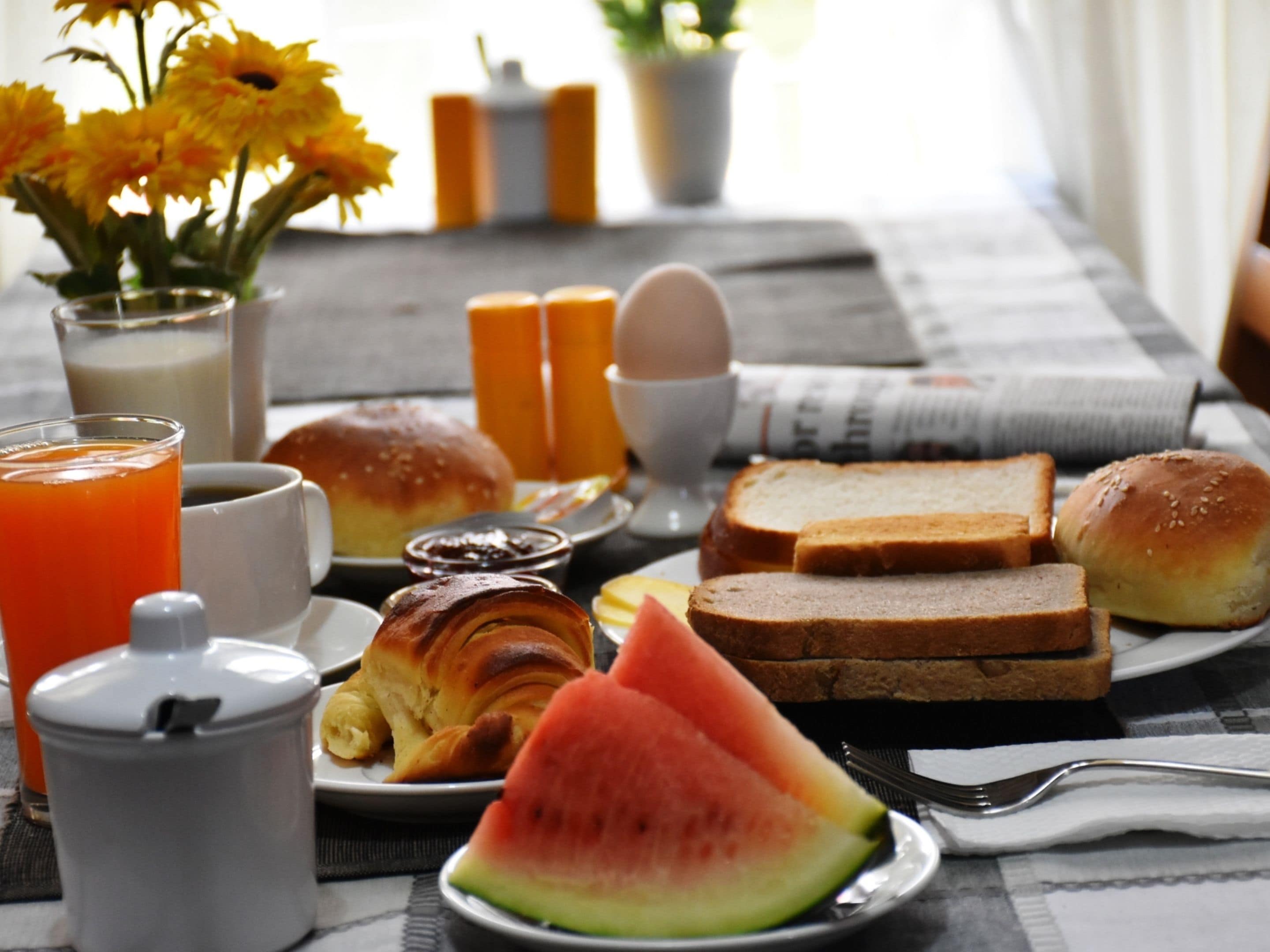 A breakfast table with croissants, bread, a slice of watermelon, a glass of orange juice, and a cup of coffee.