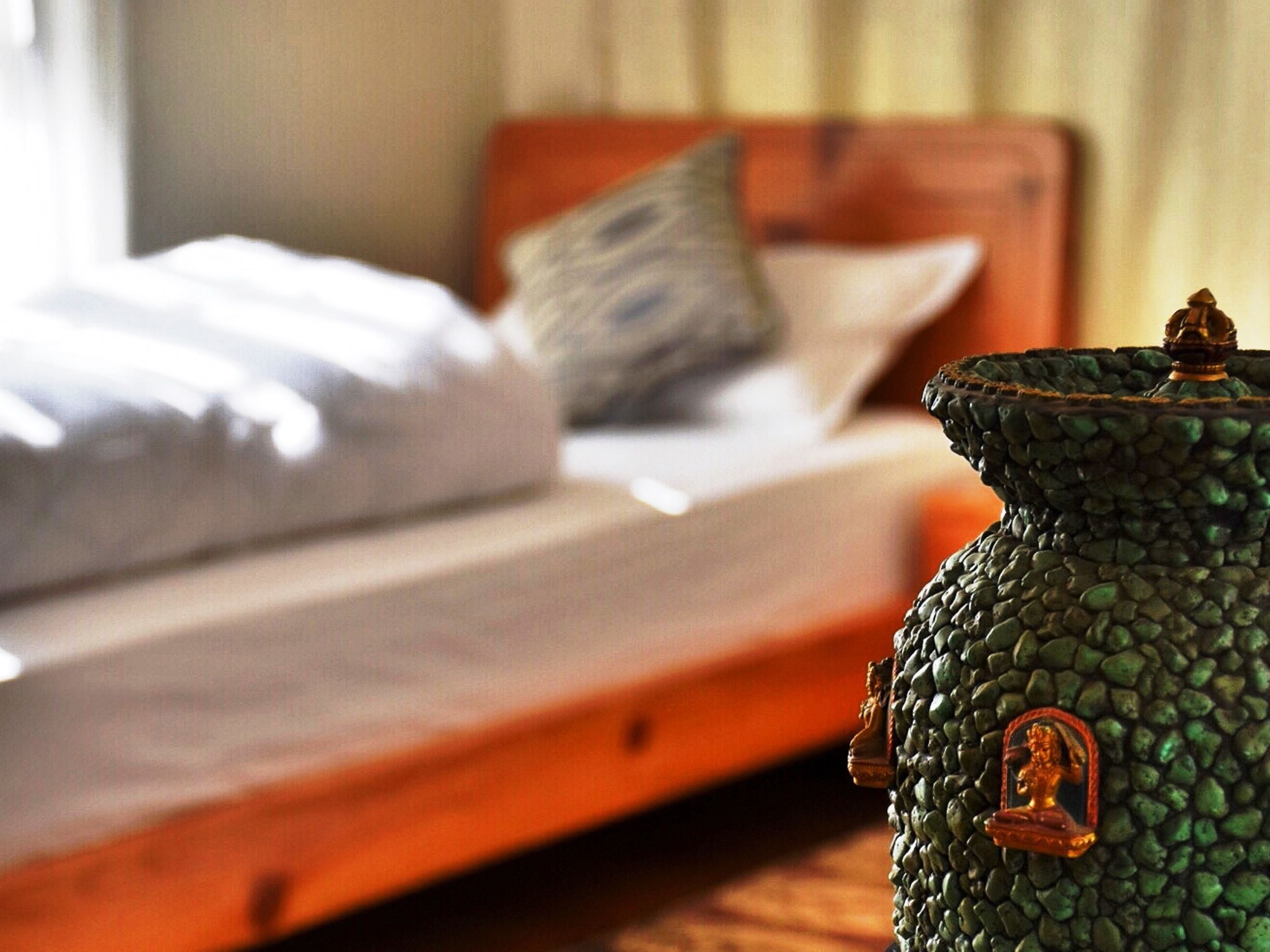 A bed with white linens and a wooden headboard, a green decorative vase with a figure on top, and a window with curtains.