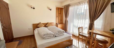A bedroom with a bed, wooden headboard, a desk with a chair, a television, and a window with curtains.