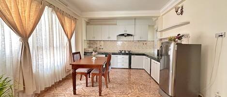 A dining area with a wooden table and chairs, a kitchen with white cabinets and a stainless steel refrigerator, and patterned floor tiles.