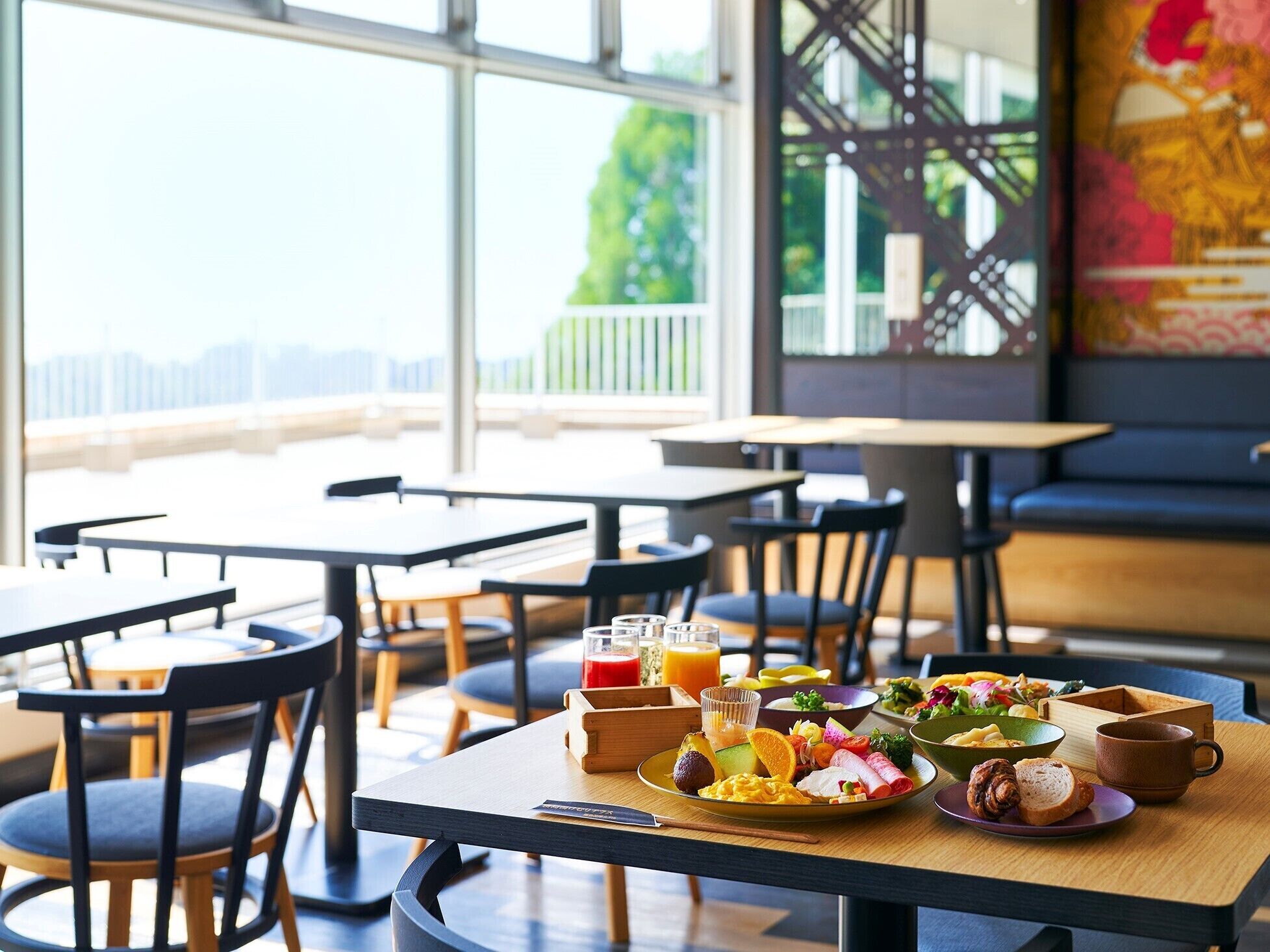 A breakfast setting with a variety of dishes, including eggs, vegetables, and a fruit bowl, on a wooden table in a bright room with large windows.