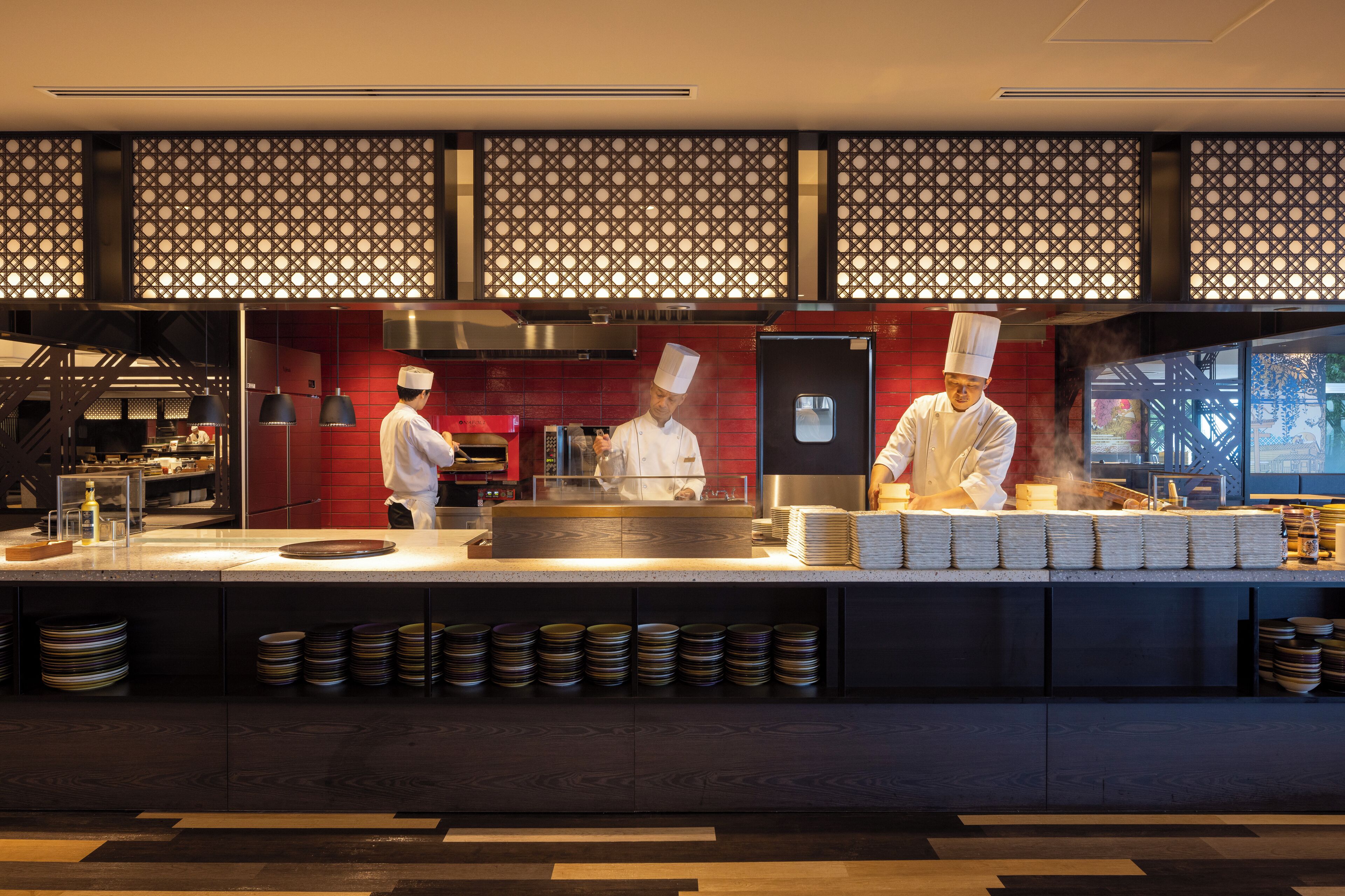 Una cocina moderna con cocineros trabajando, una isla grande con platos apilados y una pared decorativa con un diseño geométrico.