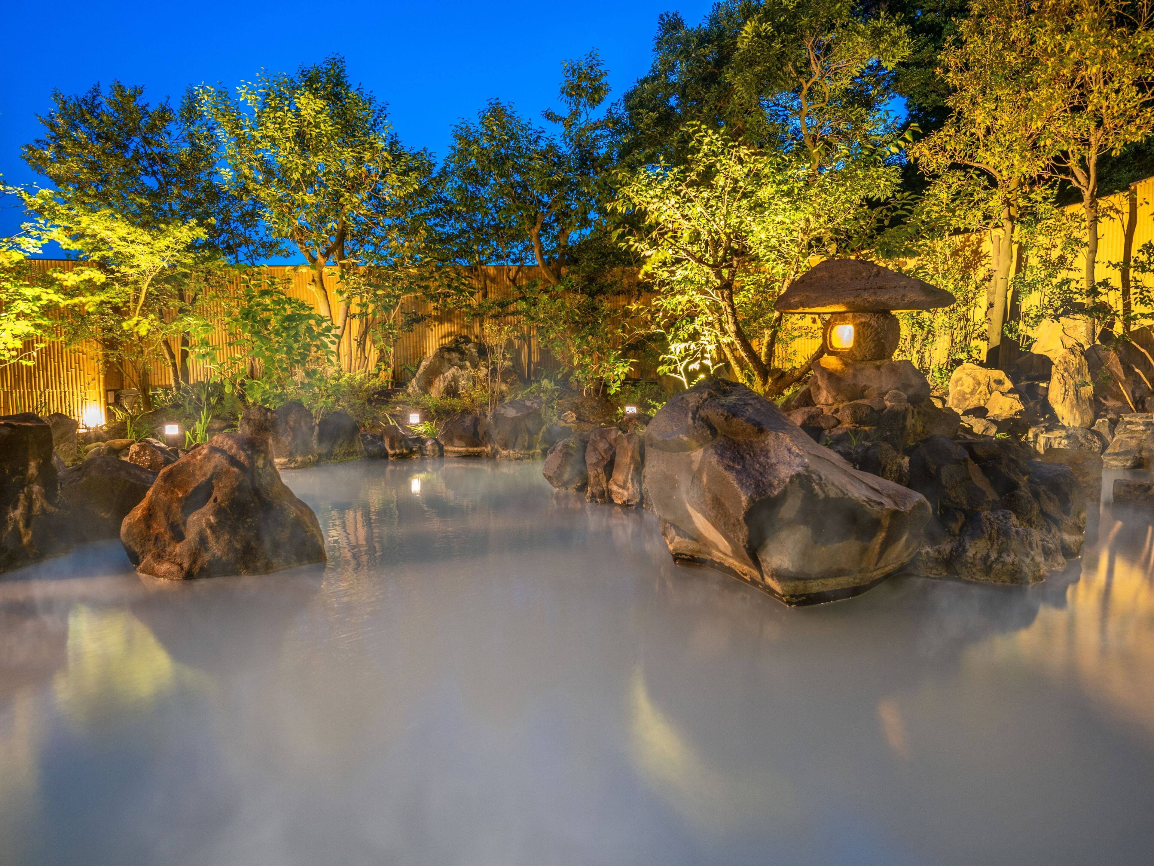 A serene hot spring area with large rocks, bamboo walls, and a traditional hut.