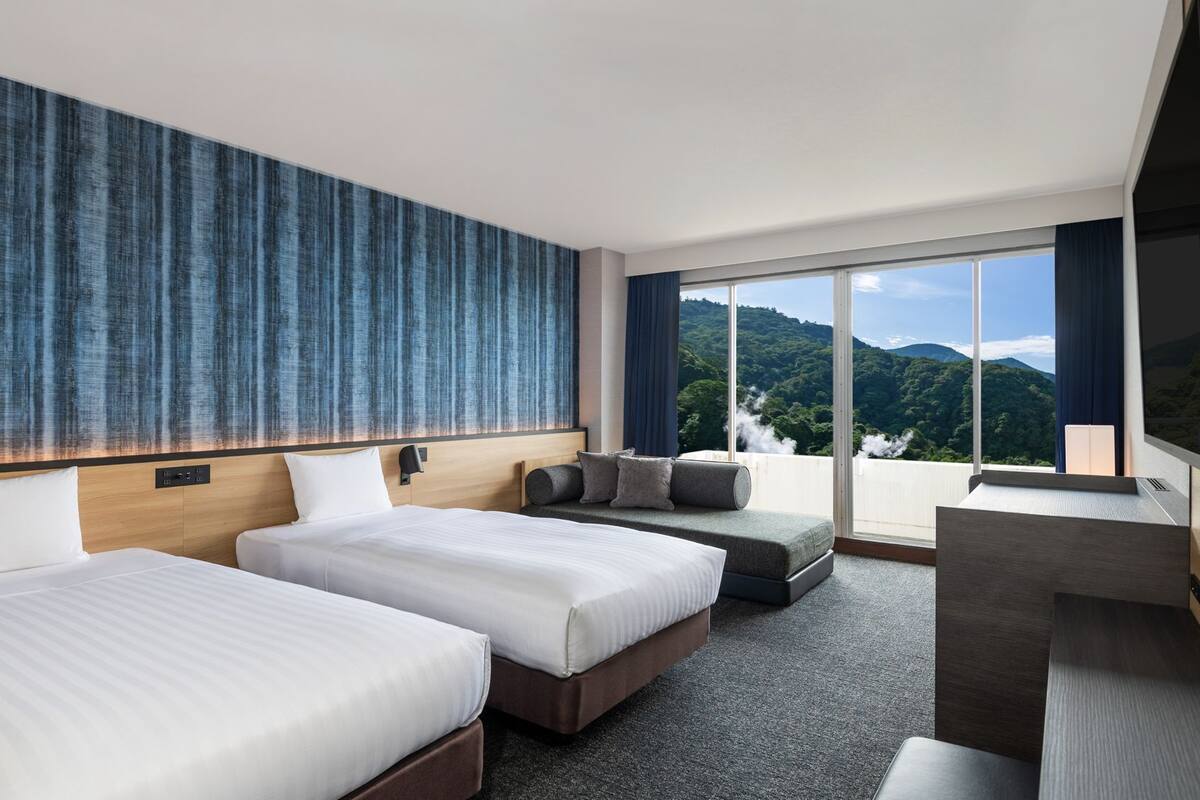 Ein Hotelzimmer mit zwei Betten, einem Sofa, einem Fernseher und einem großen Fenster mit Blick auf die Berge.