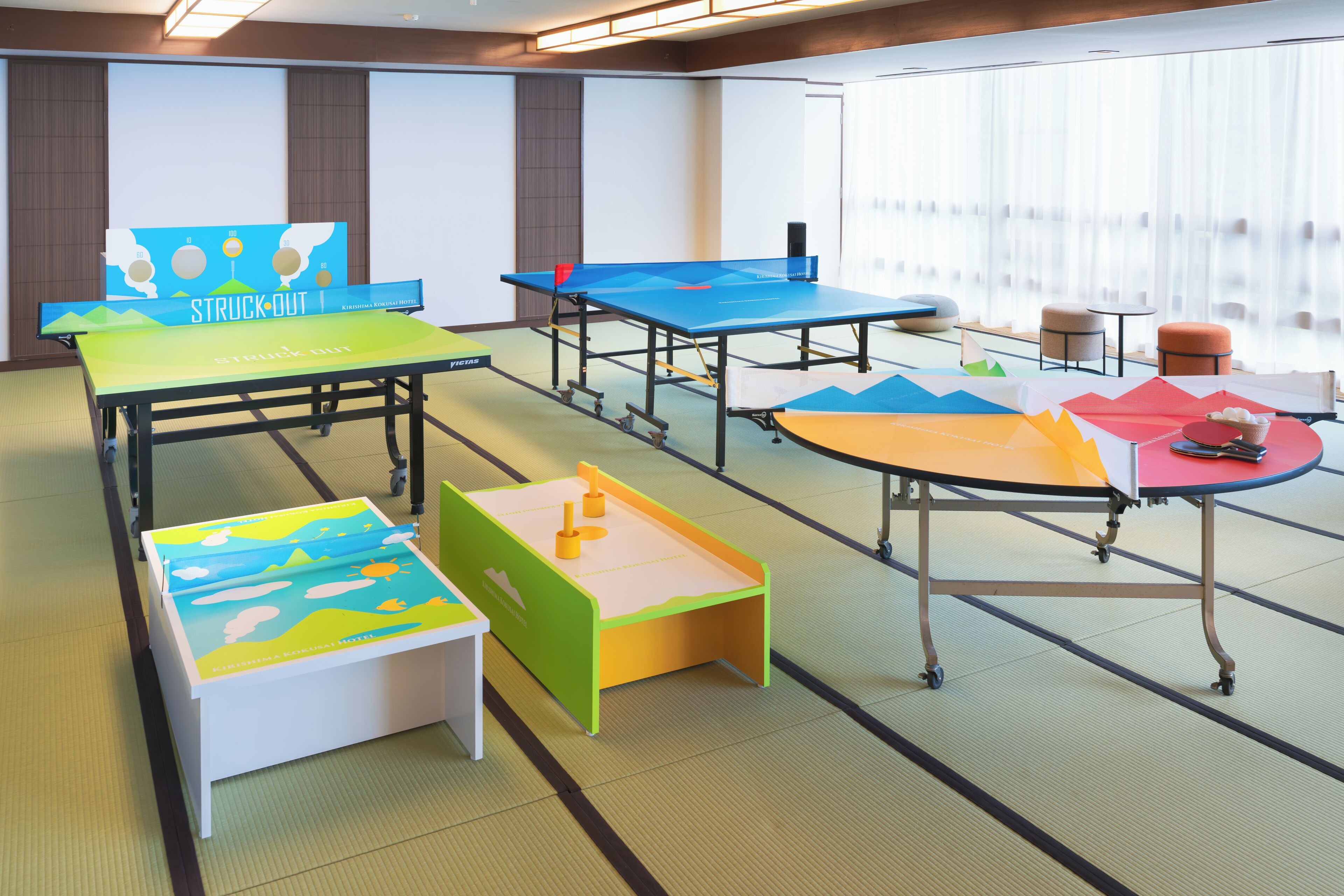 A room with tatami mat flooring, multiple ping pong tables, and a large projection screen.