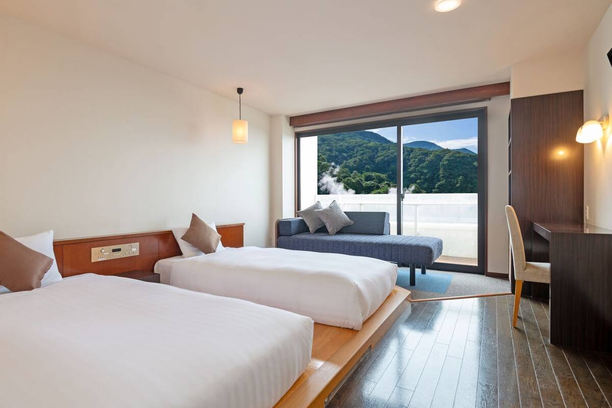 Ein Hotelzimmer mit zwei Betten, einem Sofa, einem Schreibtisch und einem großen Fenster mit Blick auf die Berge.