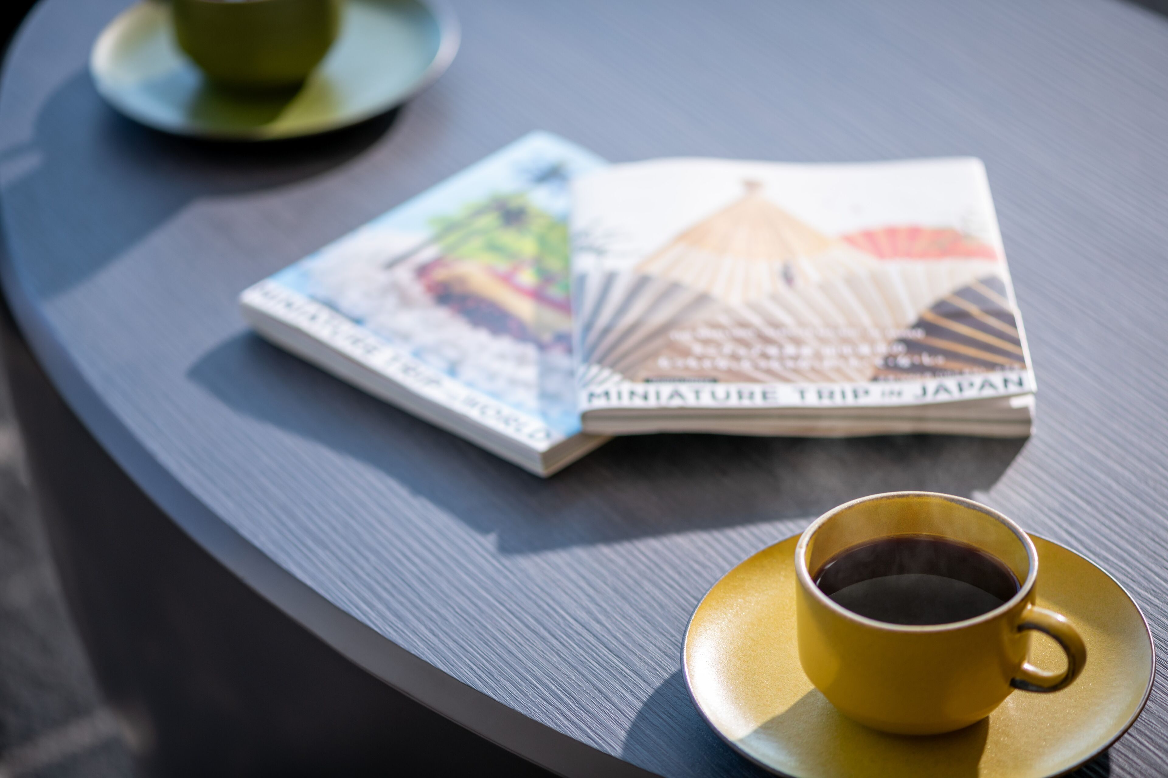 Un tavolo con una rivista dal titolo 'Miniature Trip to Japan' e una tazza di caffè su un piattino.