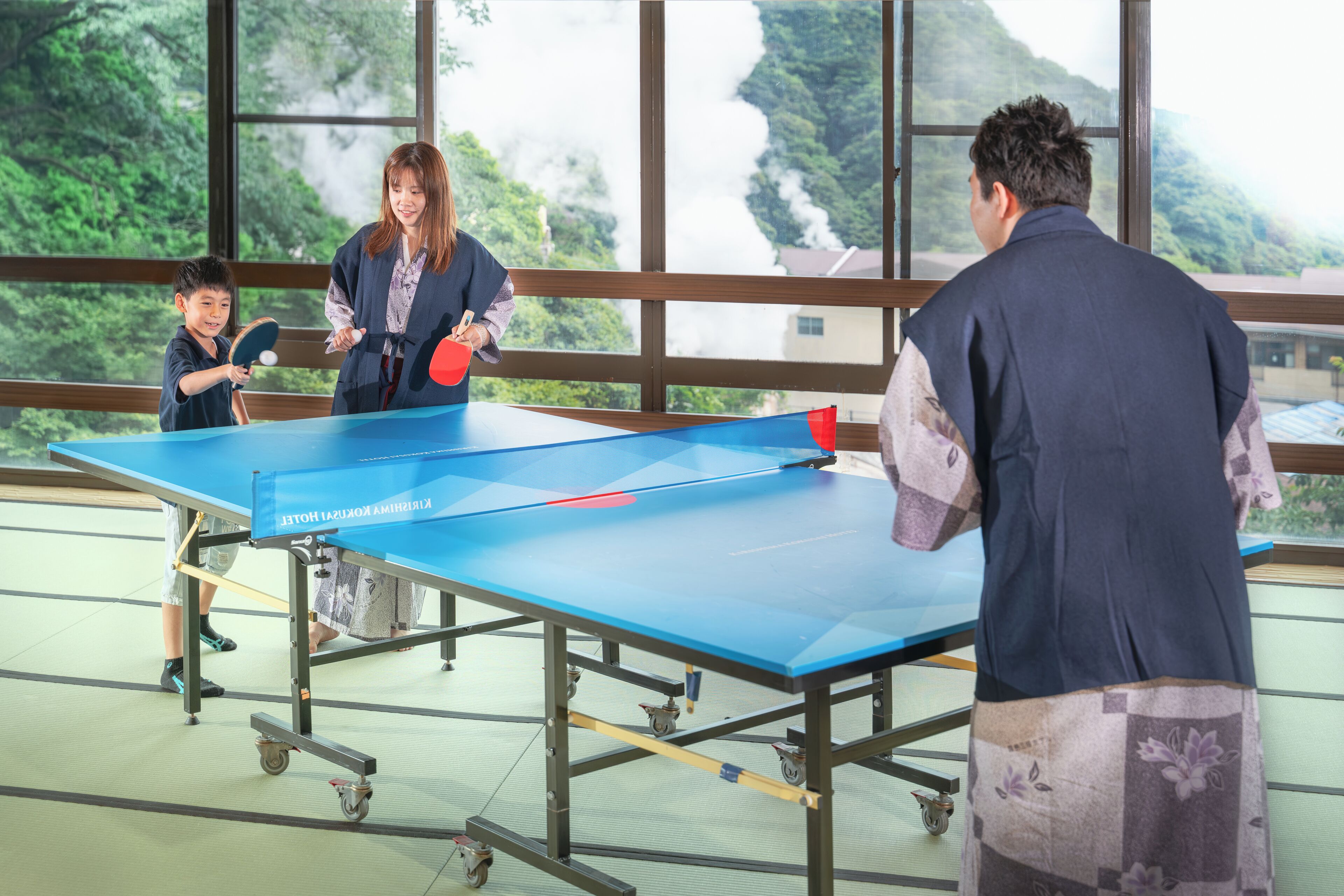 A ping pong table in a room with large windows, a view of greenery outside, and a person playing.