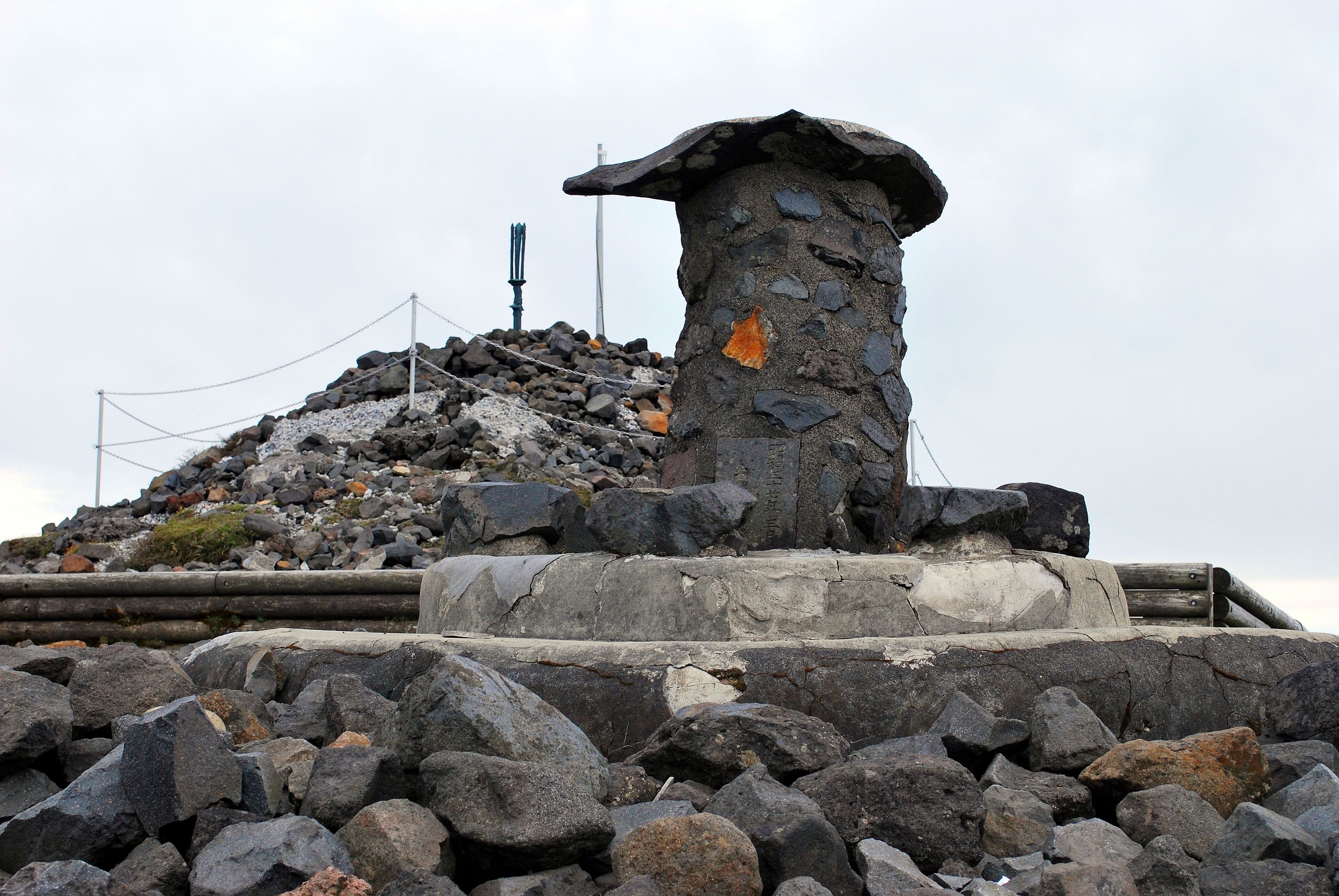 A stone monument with a weathered top, surrounded by a rocky terrain and a fence.