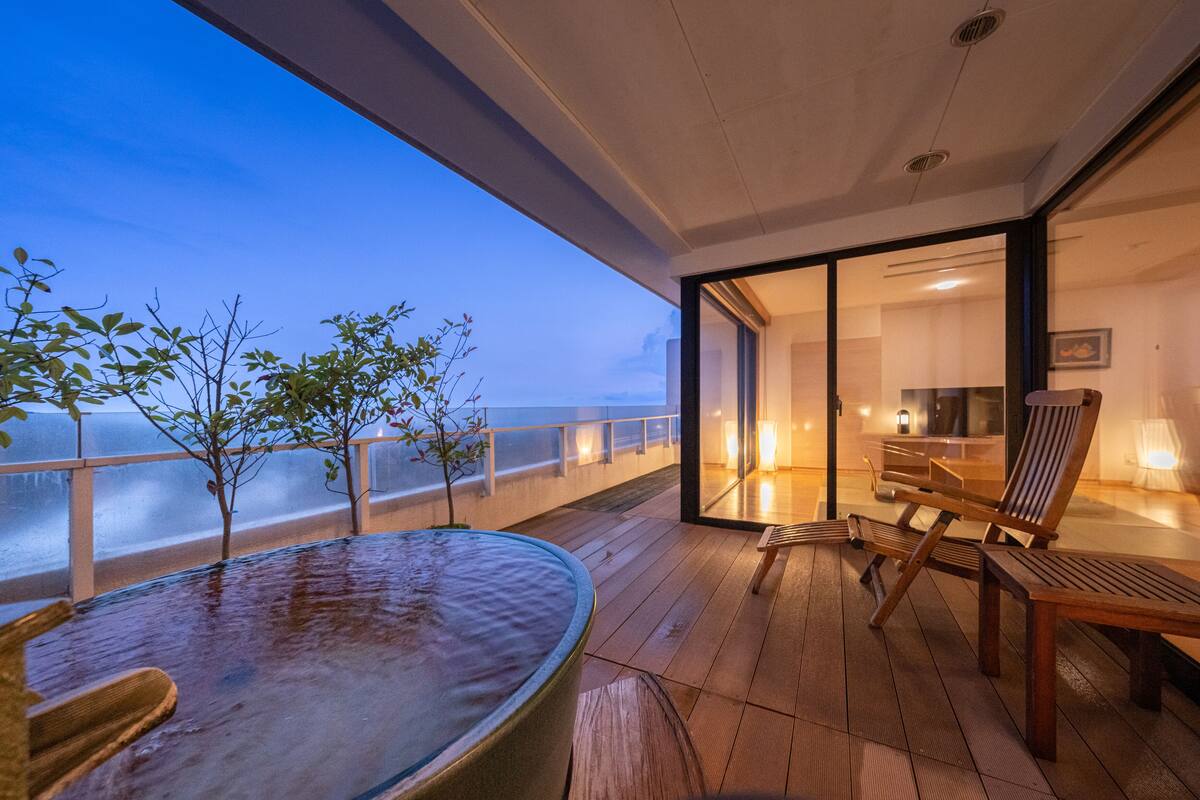 Eine Holzterrasse mit Whirlpool, Stühlen und Blick auf den Ozean im Abendlicht.