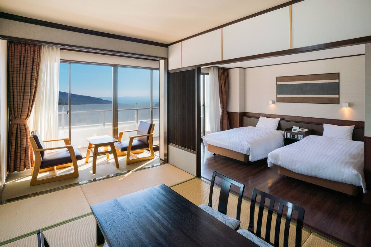 Ein Hotelzimmer mit großem Fenster, zwei Betten, einem Esstisch und Meerblick.