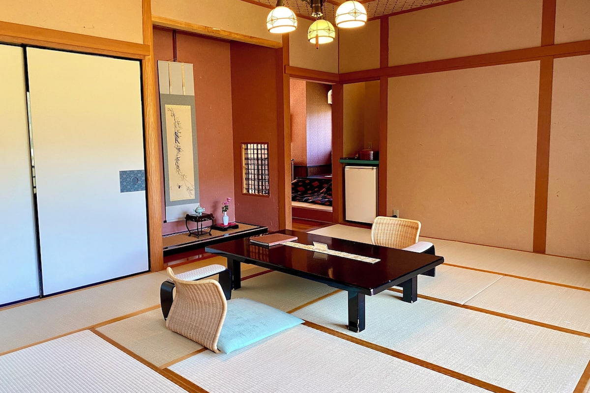 A traditional Japanese-style room with tatami flooring, a low table, a cushioned chair, a small side table with a teapot, and a refrigerator.