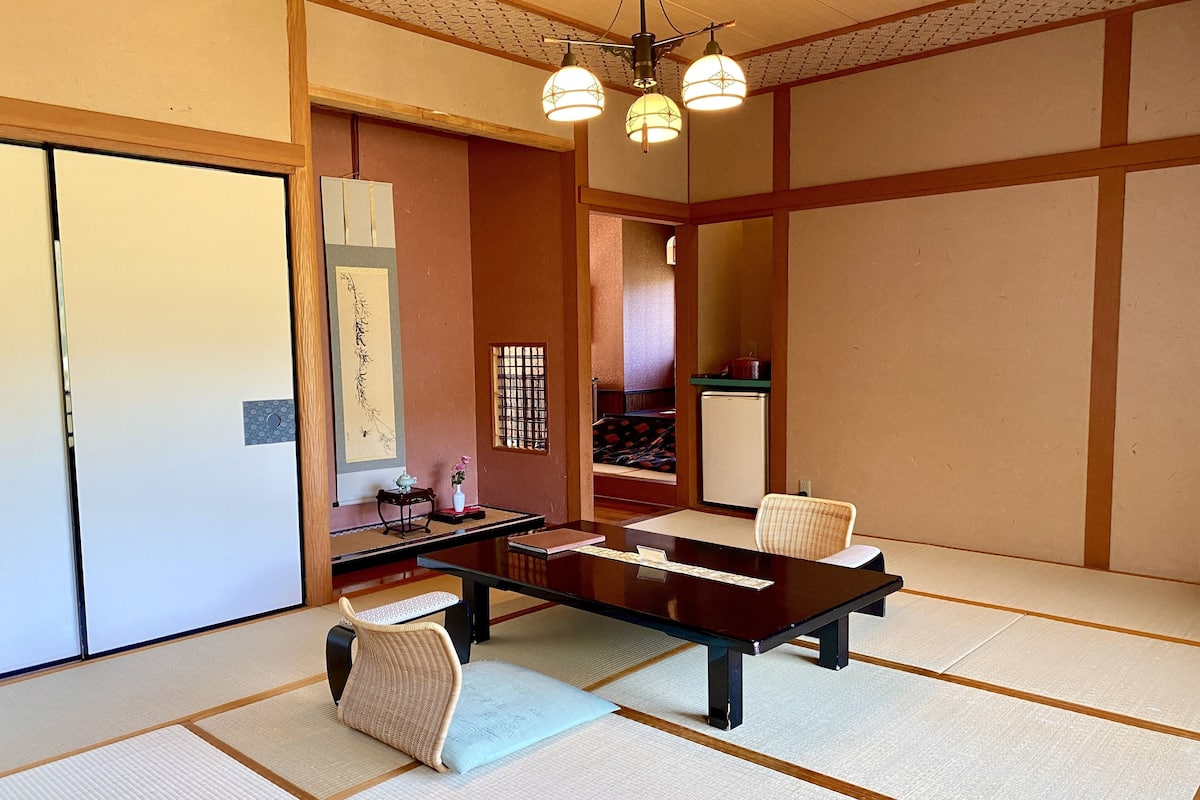 A traditional Japanese-style room with tatami flooring, a low table, a cushioned chair, a small side table with a teapot, and a refrigerator.