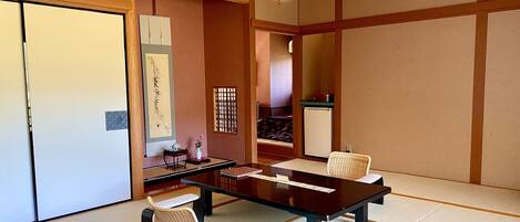 A traditional Japanese-style room with tatami flooring, a low table, a cushioned chair, a small side table with a teapot, and a refrigerator.