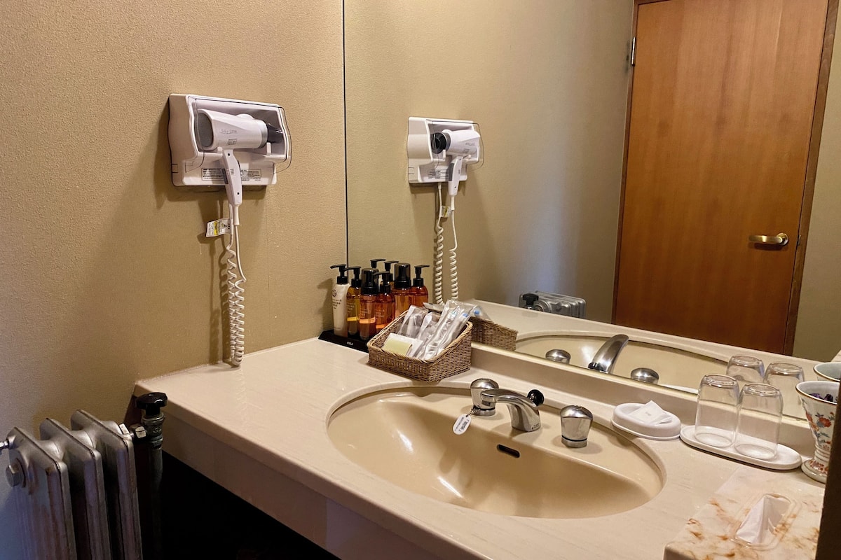 A hotel bathroom with a large sink, two hair dryers, a mirror, and a wooden door.