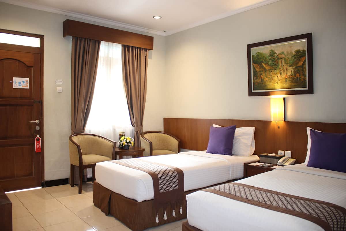 A hotel room with two beds, a wooden door, a chair, a small table with a lamp, and a painting on the wall.