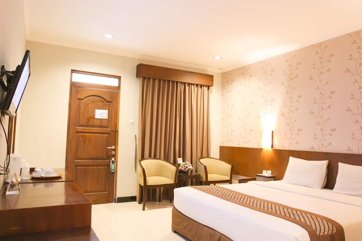 A hotel room with a bed, two chairs, a desk, a TV, and a door.