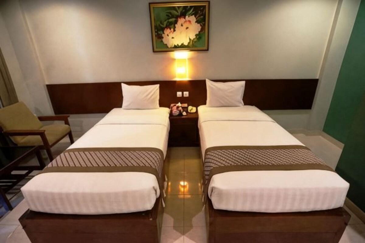 Habitación de hotel con dos camas, una mesita de noche con lámpara, una silla y un cuadro en la pared.