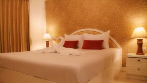 Una cama bien arreglada con sábanas blancas y almohadas rojas, dos lámparas de noche y una mesita de madera.