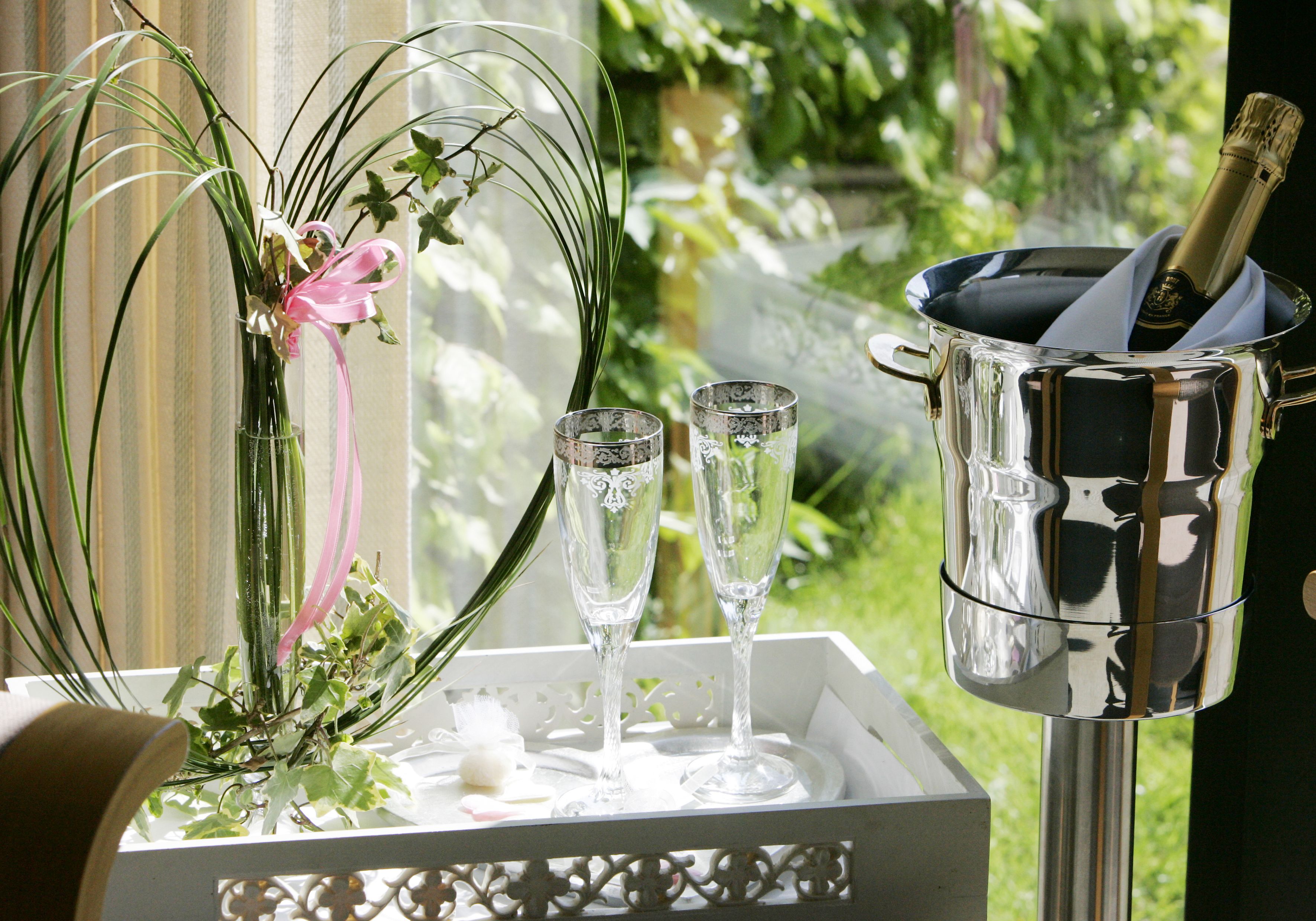 A white tray with two champagne flutes and a champagne bottle in a silver bucket, placed on a table with a floral arrangement.