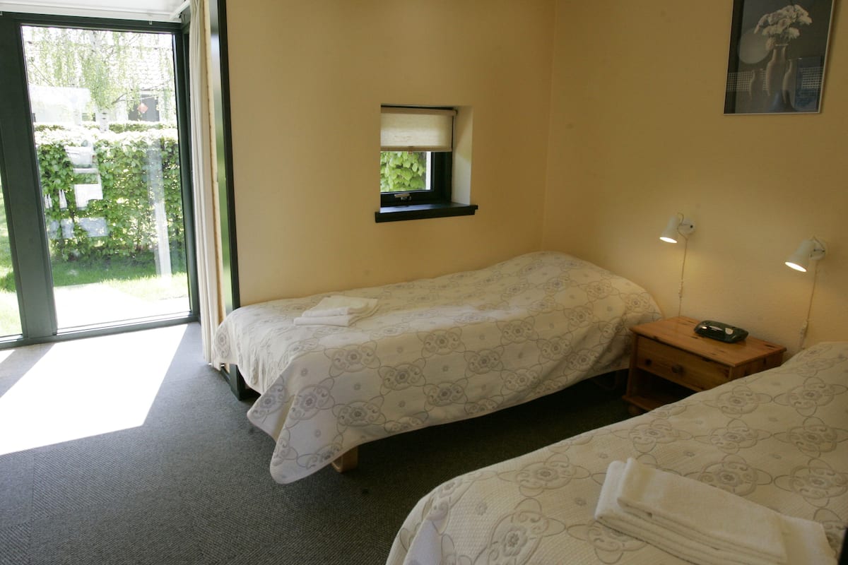 A hotel room with two single beds, a small window with a view of greenery, a bedside table with a lamp, and a wall-mounted mirror.