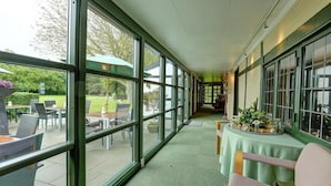 Un couloir doté de grandes fenêtres, une table recouverte d’une nappe verte et des sièges d’extérieur visibles à travers les fenêtres.