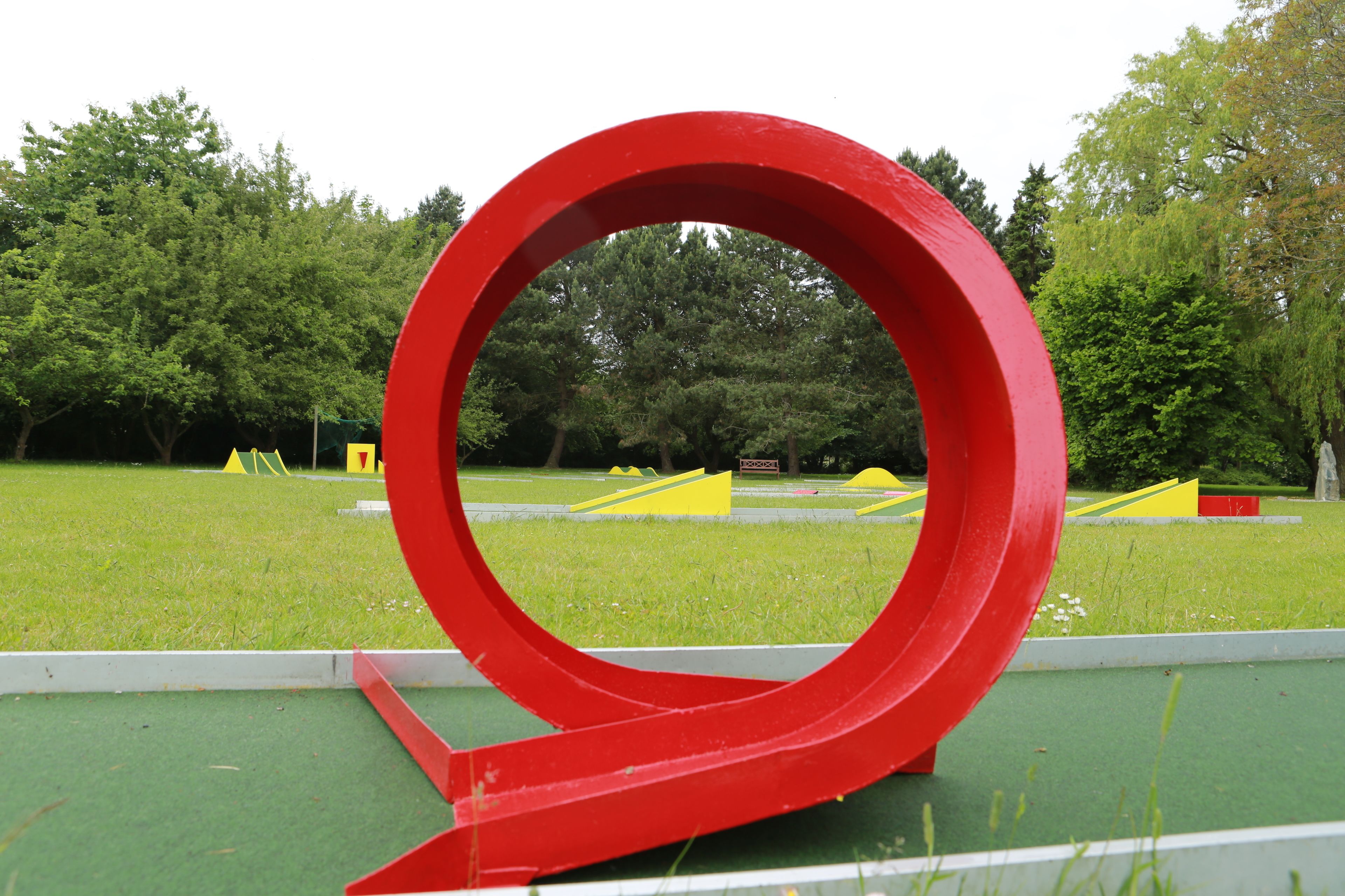 Una escultura circular roja en un parque, con césped verde y árboles al fondo.