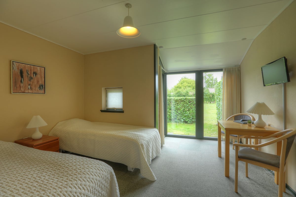 A hotel room with two beds, a desk with a chair, a television, and a window with a view of greenery.