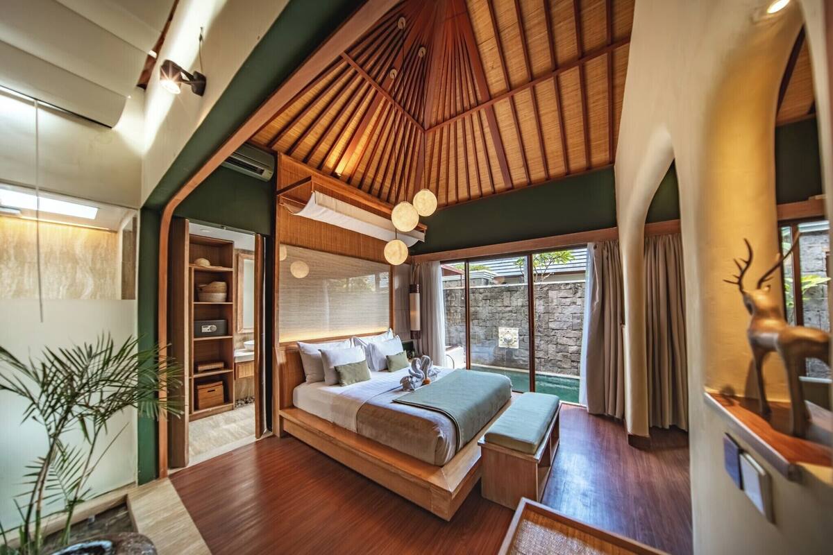 A spacious bedroom with a large bed, wooden ceiling, and a view of a stone wall outside.