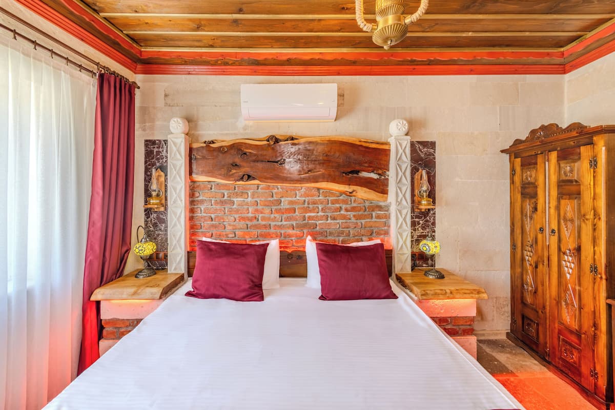 A bedroom with a brick wall, a wooden headboard, a bed with white bedding, two red pillows, a wooden nightstand with a lamp, and a wooden wardrobe.