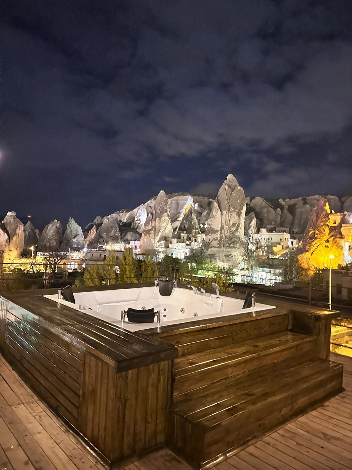 Zona de piscina en la azotea con terraza de madera, un jacuzzi y vista a un paisaje montañoso de noche.