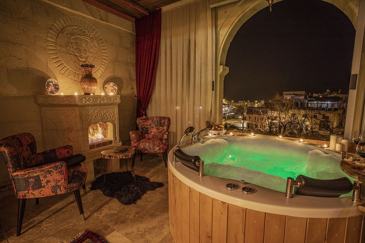 A hot tub with a view of the city at night, a fireplace, and patterned chairs.