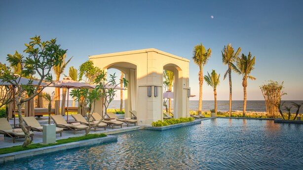 A pool area with lounge chairs and umbrellas, a covered seating area, palm trees, and a view of the ocean. - FLC Luxury Resort Samson (Sam Son)