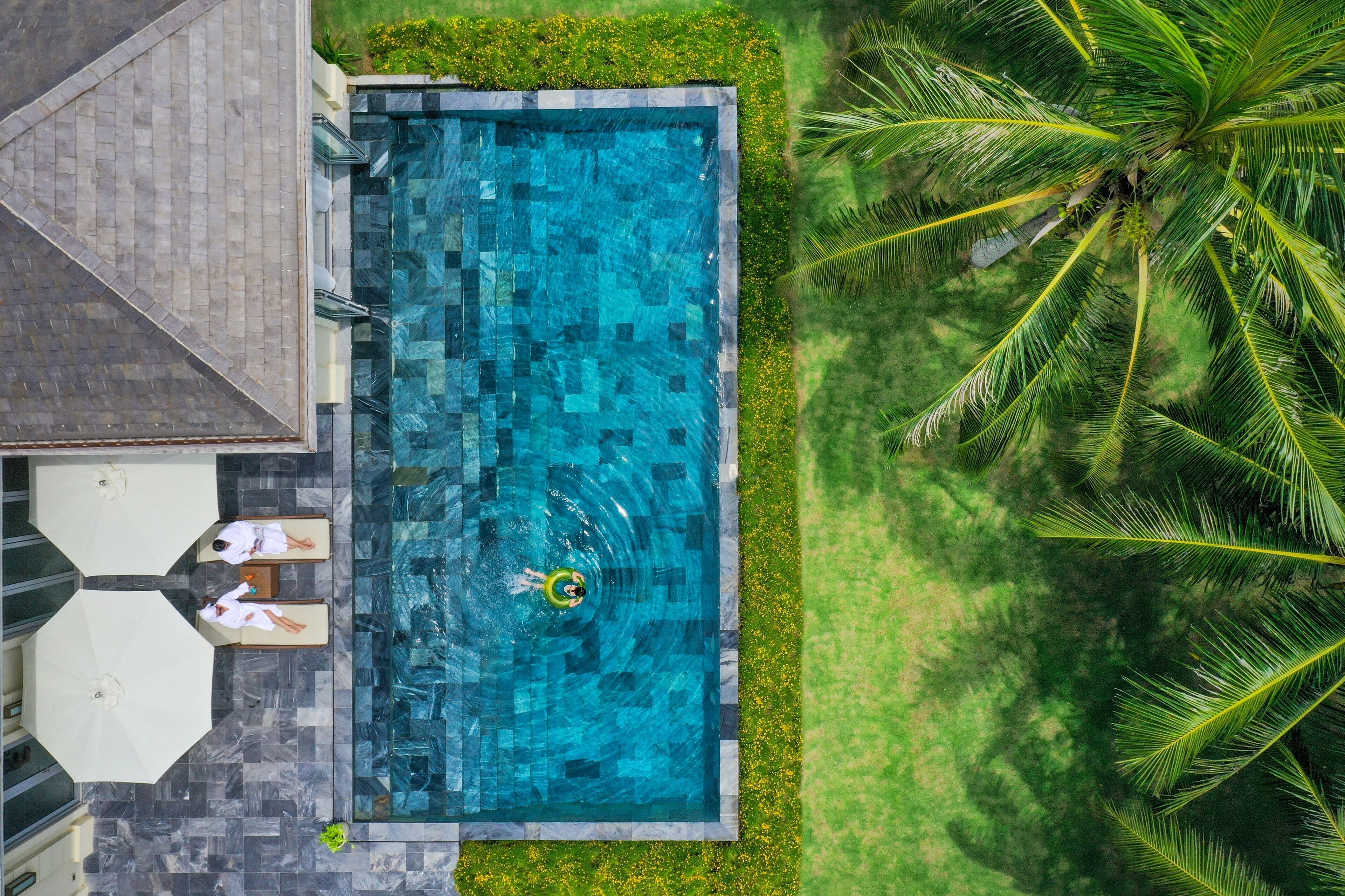 Vista aérea de una piscina rectangular con una persona en un flotador, rodeada de palmeras y un césped bien cuidado.