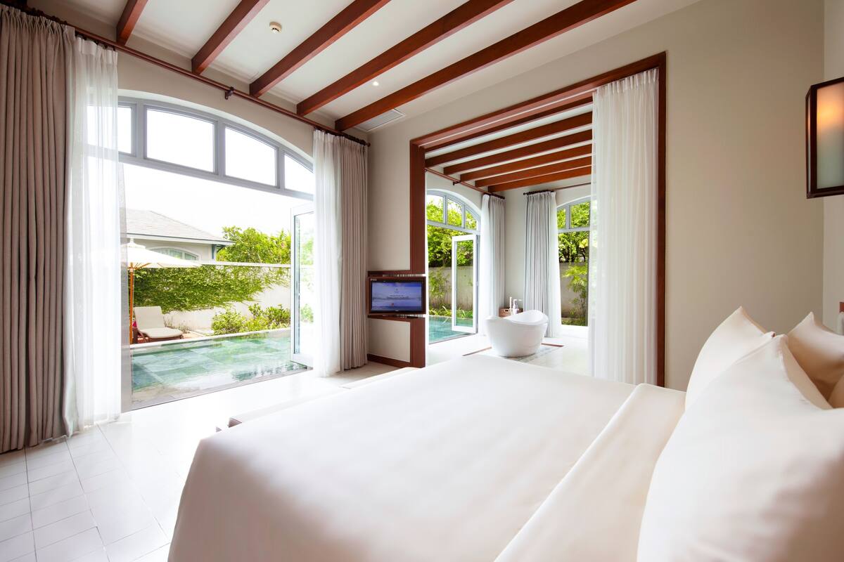 A modern hotel room with a large bed, a TV, and a view of a swimming pool and greenery.